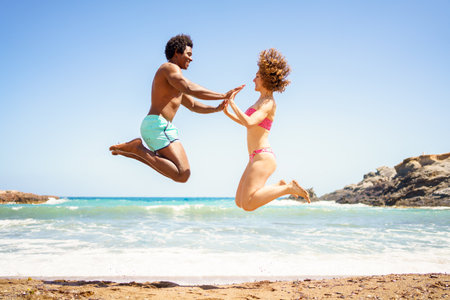 Full body side view of cheerful African American boyfriend and curly haired girlfriend, in swimwear jumping on sandy beach and holding hands near waving sea while looking at each otherの写真素材