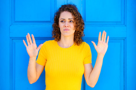 Scared young female in yellow t shirt with curly brown hair looking away with raised hands against...