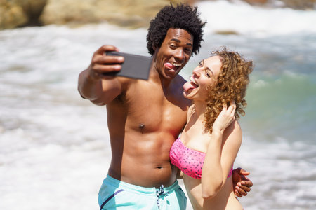 Cheerful multiracial couple in swimwear hugging and showing tongues while taking selfie on mobile phone for memory against waving oceanの写真素材