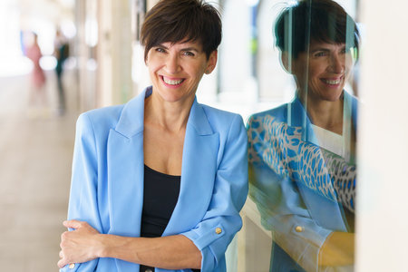 Positive middle aged female in blue jacket with short brown hair looking at camera and smiling while leaning on glass wall of modern buildingの写真素材