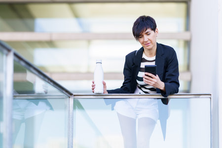 Serious entrepreneur in smart casual clothes standing near glass wall and browsing mobile phone while holding bottle of fresh waterの写真素材