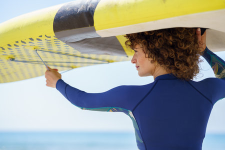 Back view of positive curly haired female in trendy swimwear with colorful paddleboard on background...