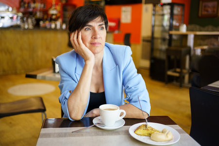 Thoughtful lady in elegant jacket sitting at table with cup of beverage and piece of cake while leaning on arm in cafeteriaの写真素材