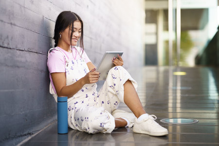 Full body glad Asian female in stylish outfit smiling and watching video on tablet while sitting on slope near thermos outside modern building in daytimeの写真素材