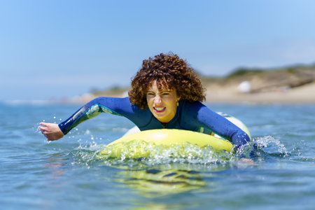 Young curly haired female surfer in wetsuit lying on SUP board and paddling with hands floating over sea against blue skyの写真素材