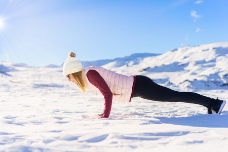 Side view of young female in warm clothes lying and doing handstand press up on snowy plain of mountainous terrain on sunny day in winterの写真素材