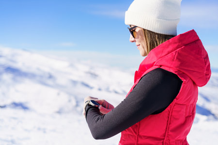 Side view of young female in warm clothes cap sunglasses, holding wristwatch and looking down to check time while standing on blurred snowy mountain slope in daylightの写真素材