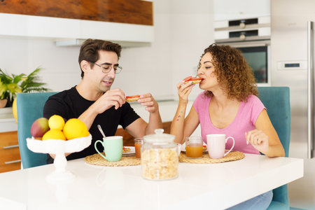 Happy adult couple in conversation and looking down while sitting at dining table in kitchen with juice in glasses, on placemat near fruits in bowl and eating salad in daylight at homeの写真素材