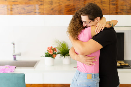 Side view of happy adult male in eyeglasses, looking down while standing in kitchen near cooking range with bread slices on grill and embracing faceless female with curly hair in daylightの写真素材