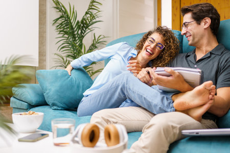 Cheerful young wife in casual clothes sitting with legs on husband, holding notebook while laughing and spending joyful moment together over couch in cozy living roomの写真素材