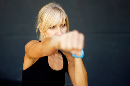 Serious young fit female boxer in black sportswear practicing punches during boxing training against gray background looking away pensively while workout at gymの写真素材