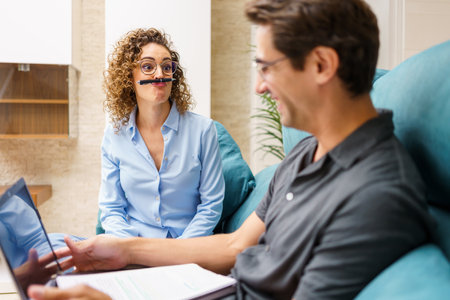 Positive young curly hair wife in smart casual shirt and eyeglasses playing with pen over mouth,...