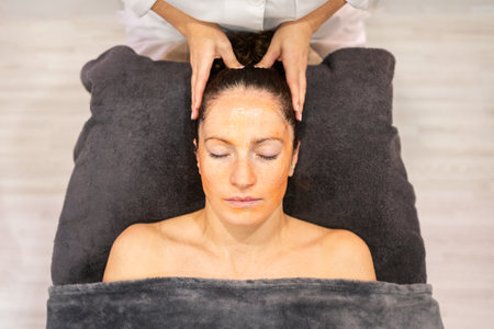 From above crop beauty specialist massaging head of female client lying on towel with closed eyes in beauty center during skin care treatmentの写真素材