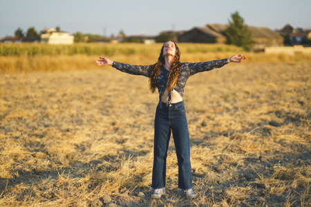 A young woman stands in a beautifully sunlit field, her arms outstretched wide as a symbol of her...