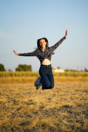 A cheerful and spirited young woman leaps joyfully and exuberantly in a sunlit golden field, embodying the essence of freedom and pure happiness against a serene and picturesque backdropの写真素材