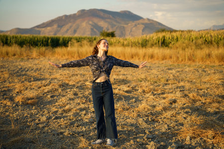 A young woman stands with her arms wide open in a beautiful golden field, completely surrounded by majestic mountains, radiating a deep sense of freedom, happiness, and pure joy in this momentの写真素材