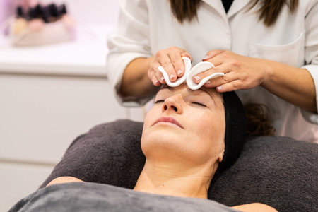 Crop unrecognizable female beautician cleaning forehead of woman using cotton pads during skin treatment in beauty salonの写真素材