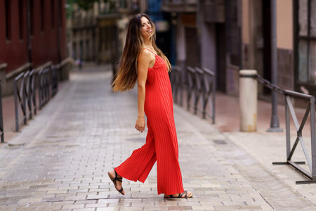 A stylish and confident woman dressed in a vibrant red dress gracefully strolls through a picturesque urban alleyway, showcasing not only her fashion sense but also her undeniable elegance and charmの写真素材