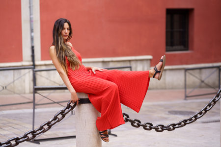 A stylish and fashionable woman dressed in a vibrant and eyecatching red dress poses playfully while leaning against a decorative chain, all set against a delightful and colorful backgroundの写真素材