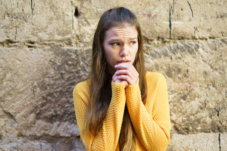 A young woman, dressed in a cozy yellow sweater, stands against an old, rustic stone wall, appearing...