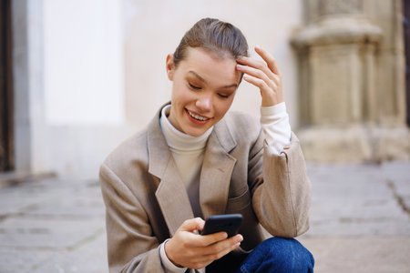 A young woman in a trendy outfit sits on the ground, happily looking at her smartphone, enjoying the...
