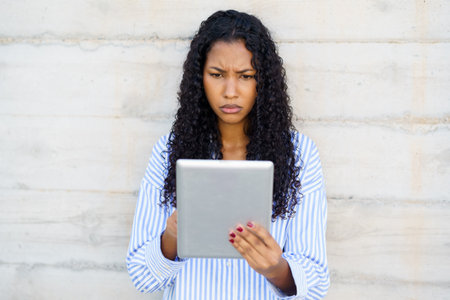 In a tranquil setting, a young woman with gorgeous curly hair is intensely focused on her sleek tablet, showcasing her strong dedication and commitment to her important tasks and knowledge pursuitの写真素材