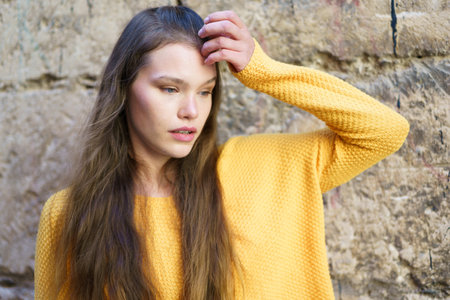 A serene young woman with flowing long hair, wearing a bright and cheerful yellow sweater, poses thoughtfully against a beautifully textured stone wall, exuding a sense of calm and eleganceの写真素材
