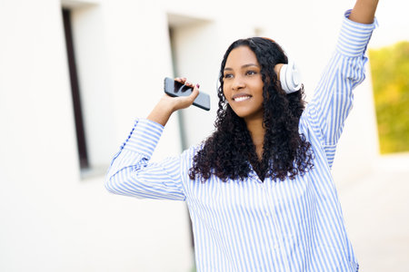 A joyful young woman dances energetically in the warm sunlight, wearing trendy headphones and skillfully holding her smartphone, truly embodying the joy of music and celebrating life in the momentの写真素材