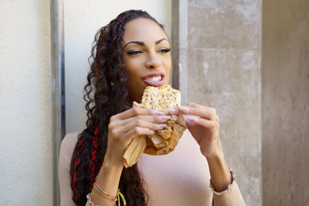 A cheerful young woman with curly hair beams with delight as she takes a satisfying bite of a...