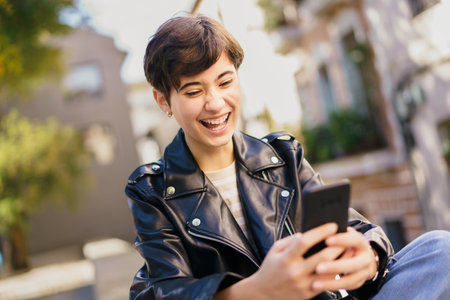 A young individual with stylish short hair and a trendy leather jacket beams with happiness as they use their smartphone outdoors, capturing a delightful moment filled with joy and deep connectionの写真素材
