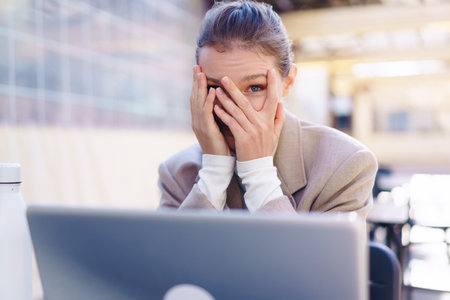 A stressed businesswoman sits at a laptop, covering her face with her hands, showing intense pressure and emotional exhaustion from her professional struggles and productivity demandsの写真素材
