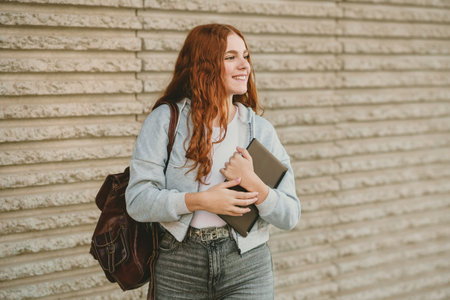 A vibrant young woman with long curly hair holds a modern tablet, wearing a trendy backpack, in front of a colorful wall, creating an inspiring atmosphere of positivity and creativityの写真素材