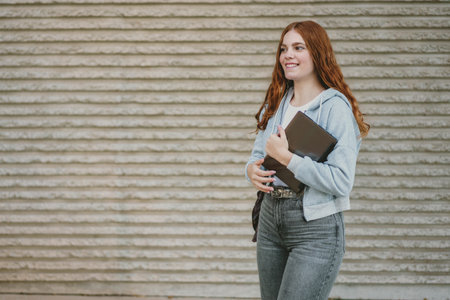 A stylish and fashionable young woman with beautiful curly red hair is smiling brightly while walking outdoors, holding a notebook, which radiates her confidence and charm effortlesslyの写真素材