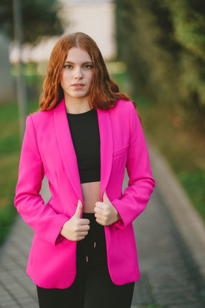 A stylish young woman confidently poses in a vibrant pink blazer and trendy black crop top, set against a beautifully blurred outdoor background that highlights her flair for modern fashionの写真素材