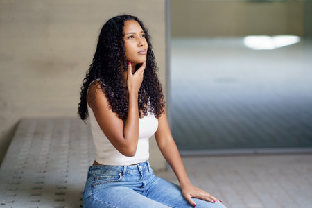 A young woman with beautiful curly hair sits thoughtfully in a bright indoor space, dressed casually...