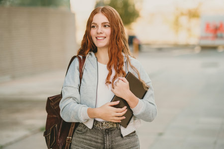 A cheerful and lively young woman with beautiful wavy red hair is happily holding several books...
