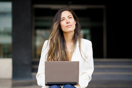 A thoughtful woman is sitting outside in a modern environment, using her laptop as she contemplates and carefully considers her next steps, decisions, and opportunities in her work and lifeの写真素材