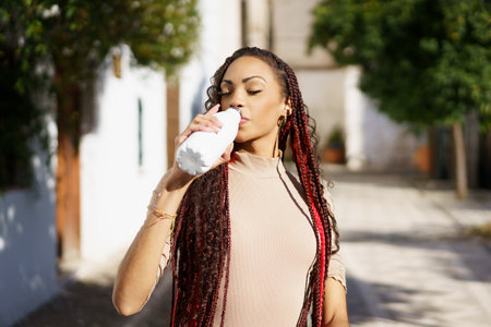 A stylish woman with long braids elegantly sips from a trendy bottle, embracing a vibrant moment in...