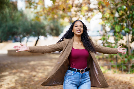 In a sunlit park with lush grass and colorful flowers, a joyful woman with curly hair spreads her arms wide, celebrating the beauty of nature and embracing the moment with freedomの写真素材