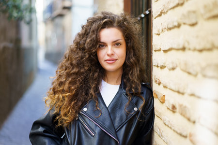 A young woman with beautifully curly hair stands confidently in a narrow and inviting alleyway, wearing an eyecatching stylish leather jacket, which enhances her charm and sense of fashionの写真素材