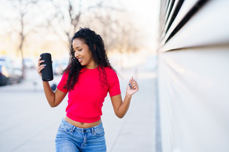A joyful woman with curly hair dances exuberantly in an urban setting. Wearing a stylish red top and trendy jeans, she radiates fun and positivity, celebrating happinessの写真素材