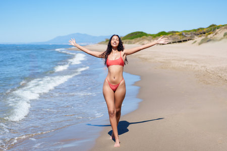 A cheerful woman wearing a vibrant pink bikini joyfully strolls along a sunny beach, embracing the gentle waves and basking in the warm sunshine, fully enjoying her time under the clear blue skyの写真素材