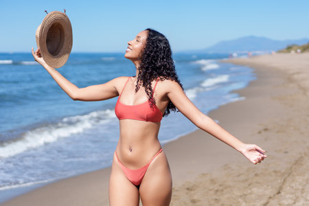 A joyful young woman poses with a stylish straw hat by the sparkling ocean, embracing the sunny beach vibe in a vibrant bikini, exuding carefree happiness and the essence of summer funの写真素材