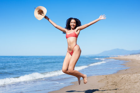 A cheerful, vibrant woman wearing a colorful bikini joyfully leaps in the air on the sandy beach, perfectly capturing the essence of summer fun and playful adventures by the ocean wavesの写真素材
