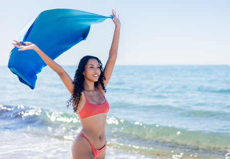 A carefree and vibrant woman revels in the sunshine on a beautiful day at the beach, joyfully dancing with her flowing blue shawl, creating a stunning silhouette against the serene ocean backdropの写真素材