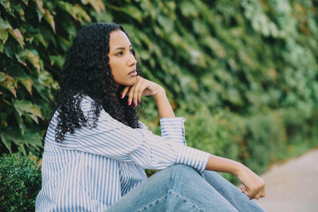 A young woman in a trendy striped shirt and stylish jeans sits outdoors, surrounded by greenery, reflecting peacefully and joyfully on the beauty of nature and enjoying the warmth of daylightの写真素材