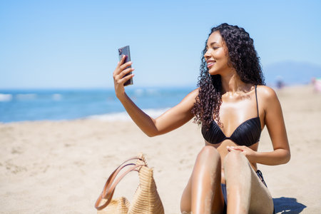 A cheerful woman, wearing a stylish black bikini, enjoys a wonderful beach day while taking a joyful selfie that perfectly captures the essence of summer vibes and her delightful, carefree lifestyleの写真素材