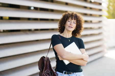 A confident young woman with curly hair stands with her arms crossed, holding a notebook. Shes casually dressed and smiling softly against a striking modern architectural backgroundの写真素材