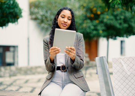 A fashionable woman is sitting outdoors, absorbed in her tablet, all while being surrounded by an...