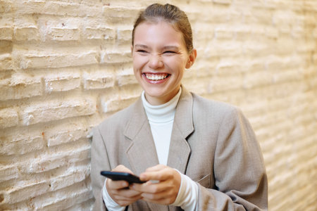 A cheerful young woman in a stylish blazer smiles widely as she engages with her smartphone against...